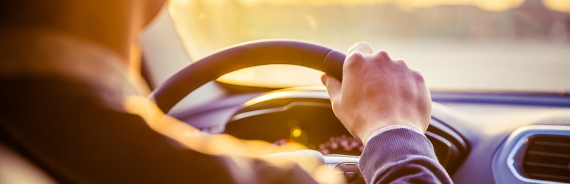 A person drives a car during sunset, viewed from the back seat with sunlight streaming through the windshield and the dashboard softly illuminated.